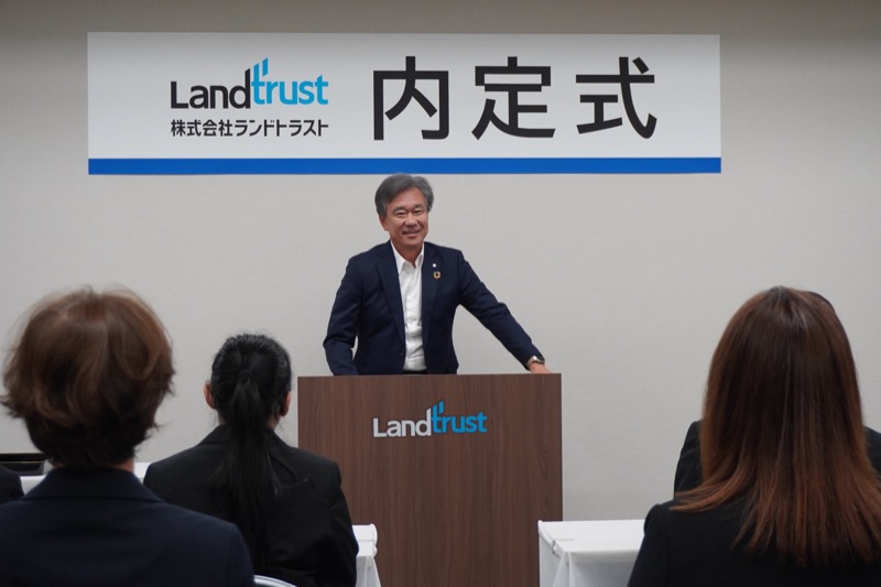 「内定式」の看板を背景に、笑顔で挨拶する社長